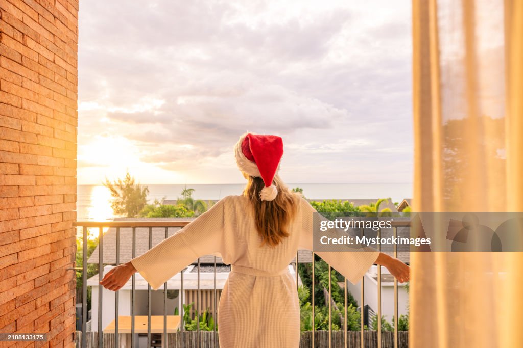 Woman in robe standing on balcony with open arms at sunrise.