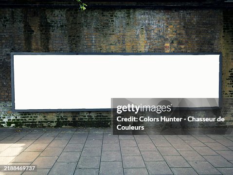 A long, empty, white, rectangular billboard in front of a weathered bricks wall. Paved sidewalk. Sunlight. Natural colors. A long, empty, white, rectangular billboard in front of a weathered bricks wall. Paved sidewalk. Sunlight. Natural colors.