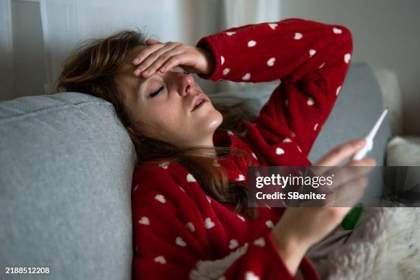 sick woman feeling unwell checking her temperature on sofa - temperatura fotografías e imágenes de stock