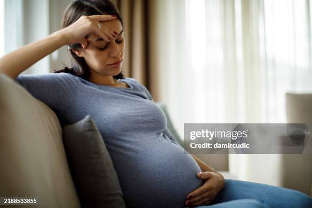 müde, gestresste schwangere, die auf einer couch sitzt und sich den bauch hält - muskelkontraktion stock-fotos und bilder