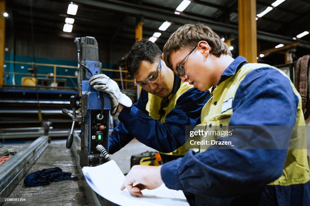 Two workers in safety glasses and vests operate machinery in a factory, focusing on a blueprint as they collaborate on a project