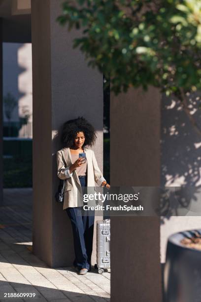 professional woman traveling outdoors with luggage and smartphone in sunlight - travel essentials stock pictures, royalty-free photos & images