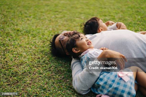 a asian adult father lies on the grass with his two young children, enjoying a peaceful moment outdoors in a natural setting. - eenjarig plantenkenmerk stockfoto's en -beelden