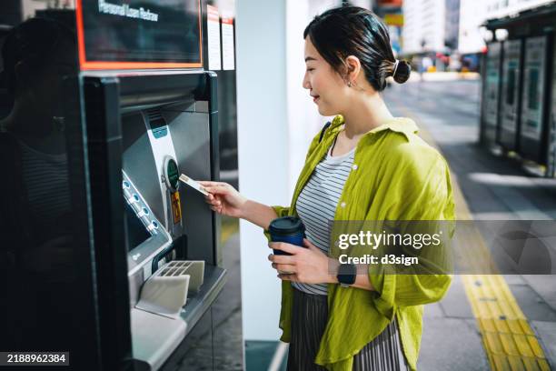 young asian woman inserting bank card into automatic cash machine in city street. withdraw money, pay bills, check account balances and make a bank transfer. personal banking. privacy protection. mobile banking security concept. smart banking on the go - geldautomat stock-fotos und bilder