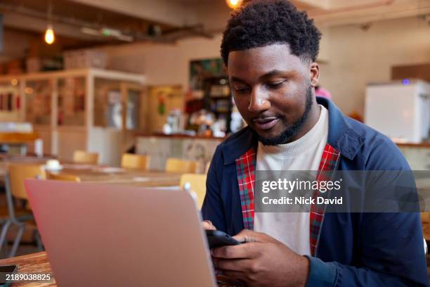 a young man text messaging on his mobile phone while working in a local cafe - hot desking stock pictures, royalty-free photos & images