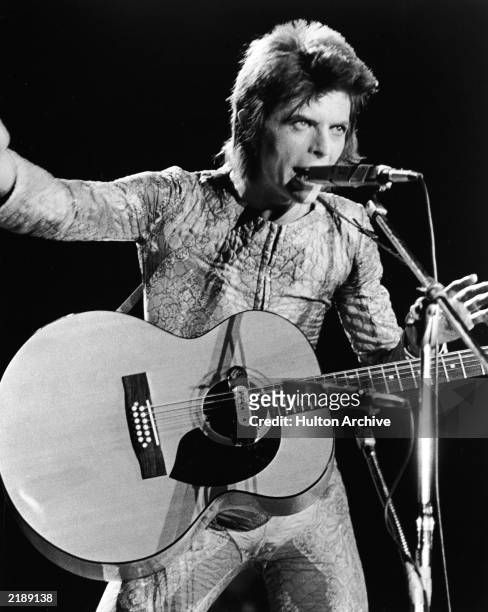 British rock singer David Bowie performs with an acoustic guitar on stage, in costume as 'Ziggy Stardust,' circa 1973.