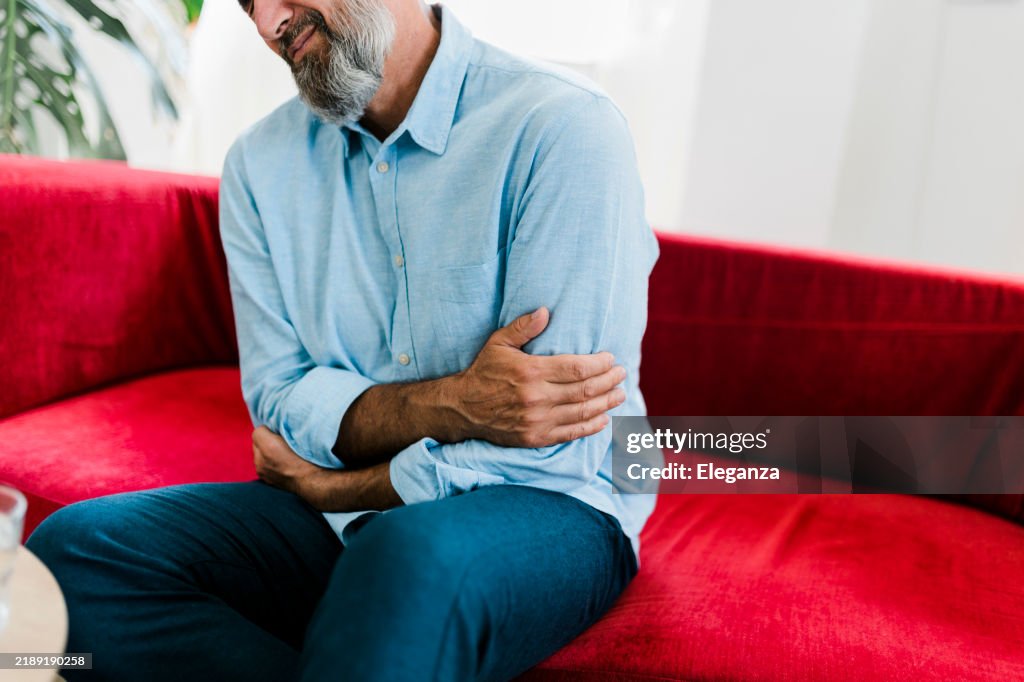 Senior man feeling unwell, suffering from backache, massaging aching muscles while sitting on sofa in the living room at home. Elderly and health issues concept