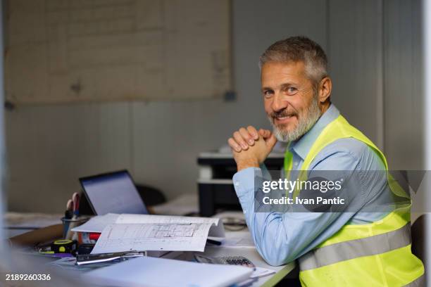 selbstbewusster bauarbeiter, der am schreibtisch mit blaupausen und laptop lächelt - projektmanagement stock-fotos und bilder