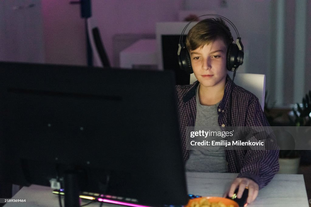 Competitive teenage boy, video game player with headphones playing video game at dark room at home. His face is illuminated by neon, RGB light of computer screen. Children gaming addiction.