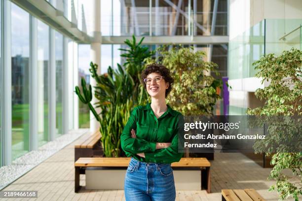 businesswoman standing with crossed arms and smiling in a sustainable office with large windows - objetivos de desenvolvimento sustentável imagens e fotografias de stock