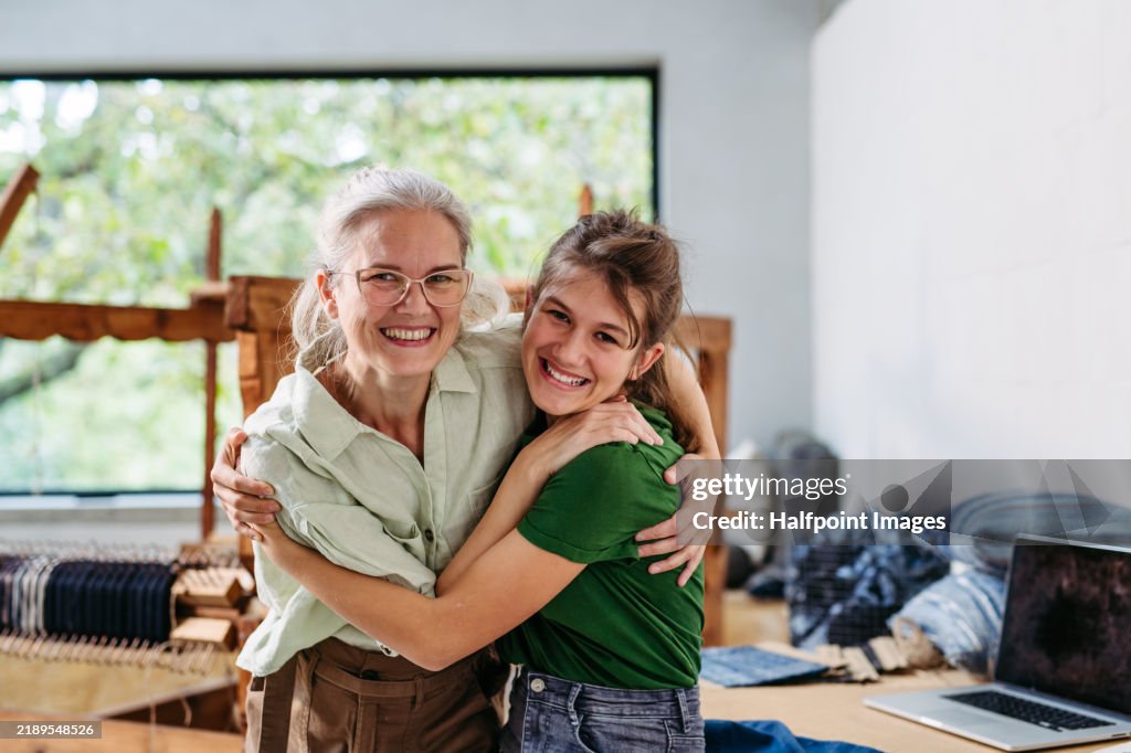 Portrait of mother and daughter in family workshop, hugging. Sustainable business, using recycled materials in fashion.