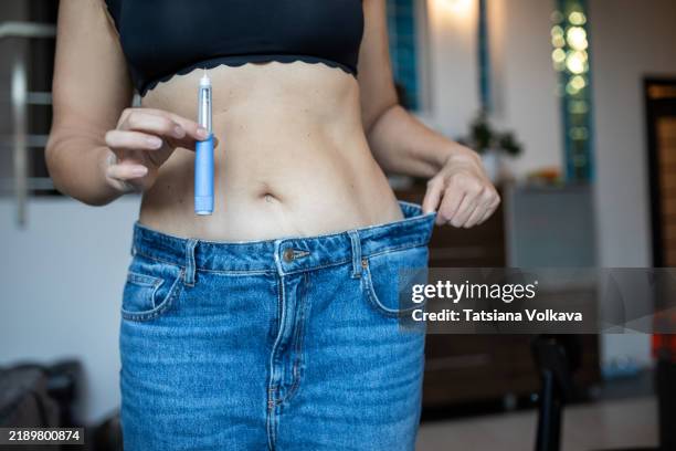 demonstrating weight-loss success, a woman shows loose jeans while holding a semaglutide injection device. - schlank stock-fotos und bilder