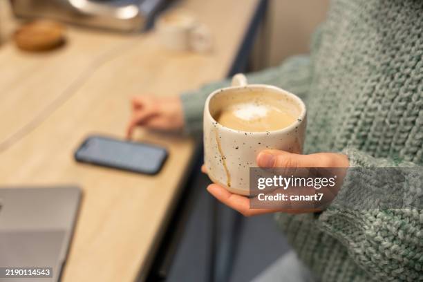 woman using smartphone and holding a cup of coffee at home office desk - koffiekop stockfoto's en -beelden