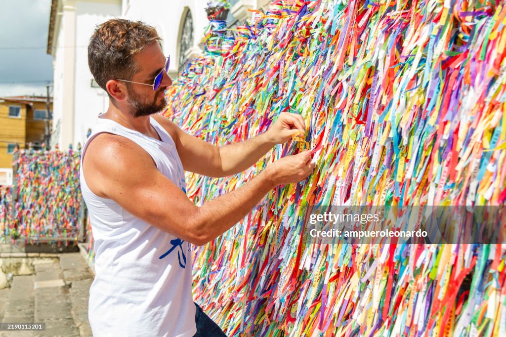 Weißer lateinamerikanischer männlicher Backpacker-Tourist, der buntes Band an Kirchenwand in Nosso Senhor do Bonfim, Salvador de Bahia, Bahia, Brasilien bindet.