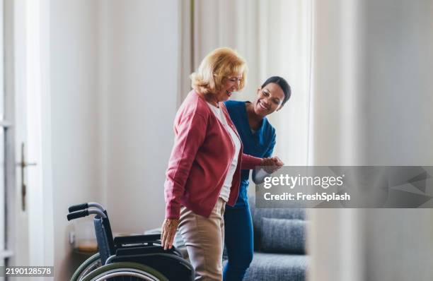 caring nurse assisting elderly woman with mobility near wheelchair in cozy living room - maatschappelijke dienstverlening stockfoto's en -beelden