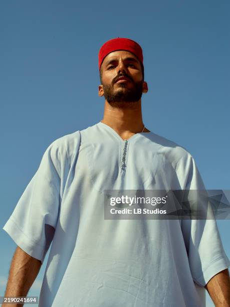 confident man with a beard wearing a traditional white robe and red headband against a clear blue sky, morocco - traditional clothing stock pictures, royalty-free photos & images