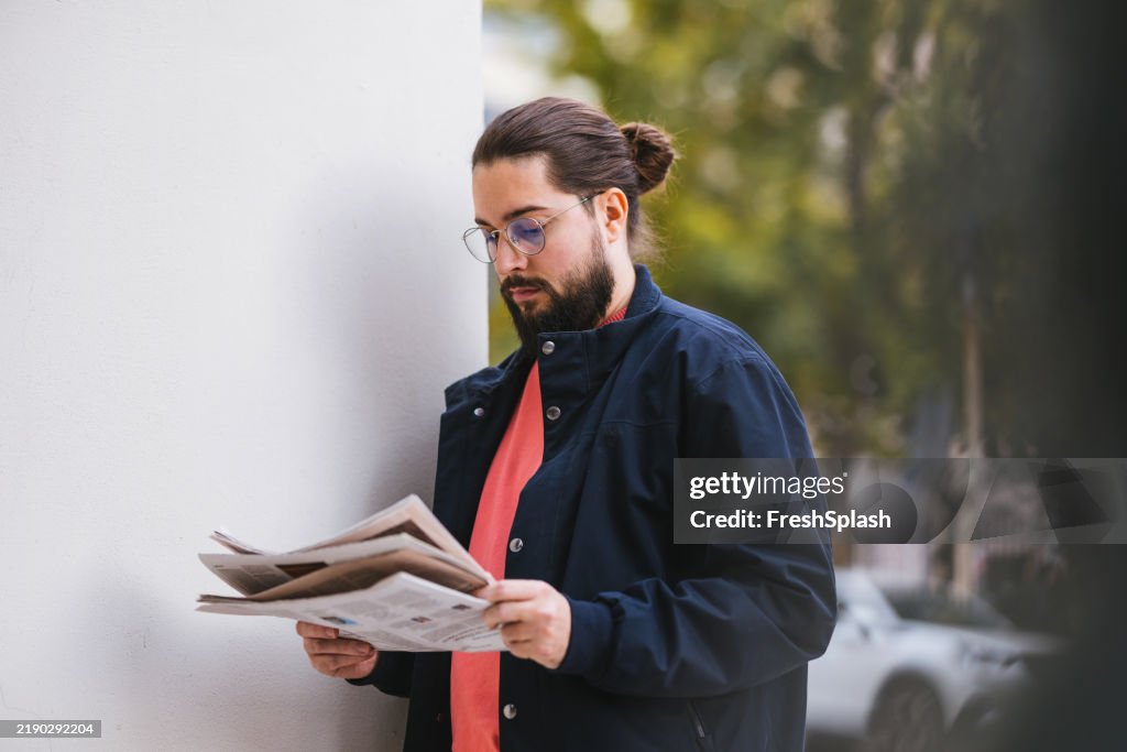 Uomo che legge un giornale all'aperto in abbigliamento casual vicino a un muro bianco