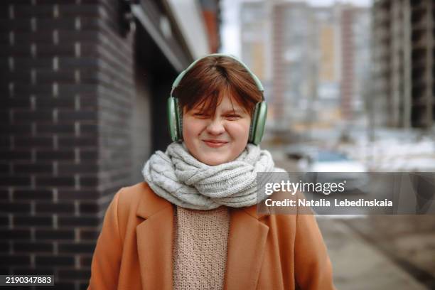 funny stylish teen girl in a scarf and big headphones, walking through a wintery city while listening to music. - kort haar stockfoto's en -beelden