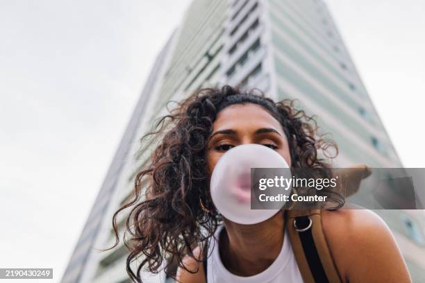 woman blowing bubble gum bubble in front of residential building - kauwgom stockfoto's en -beelden