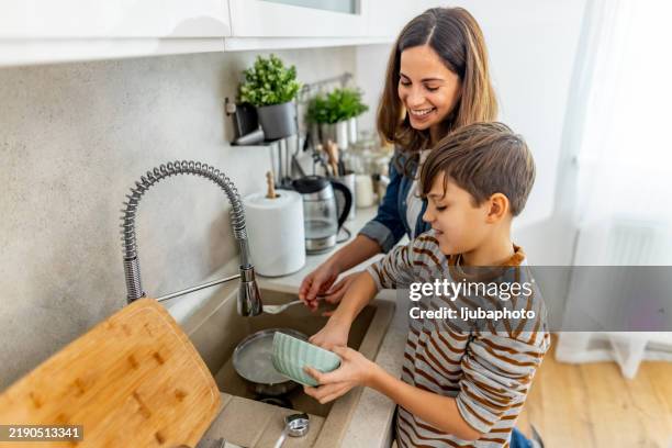 mother and son smiling and washing dishes together in a modern kitchen, sharing a moment of domestic life - afazeres domésticos imagens e fotografias de stock