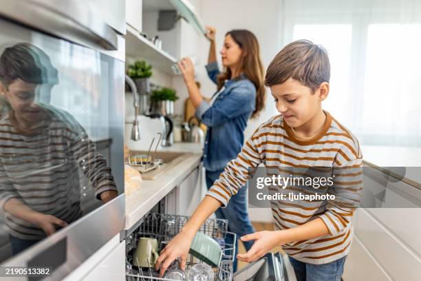 madre ayudando a su hijo a descargar el lavavajillas, compartiendo las tareas domésticas en una cocina elegante, fomentando el trabajo en equipo y las habilidades para la vida - tarea doméstica fotografías e imágenes de stock