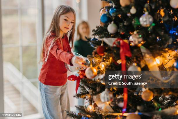 young girl decorating a christmas tree with ornaments inside a festive home - embellishment stock pictures, royalty-free photos & images