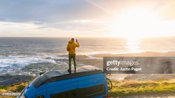 a mature van stands on the roof of his campervan as the sun rises over the north sea. he take a photo on his smartphone - north sea stock pictures, royalty-free photos & images