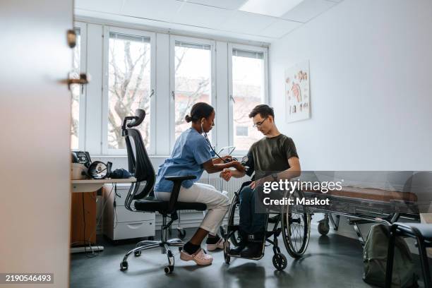 female doctor checking blood pressure of teenage boy sitting on wheelchair at examination room at clinic - orthopädisches hilfsmittel stock-fotos und bilder
