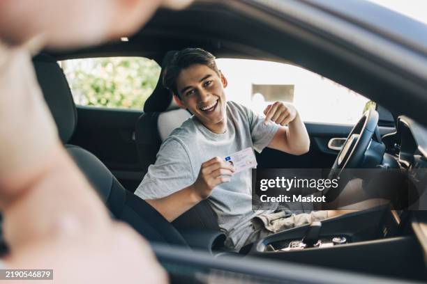 happy young man showing drivers license while sitting in car - patente di guida foto e immagini stock
