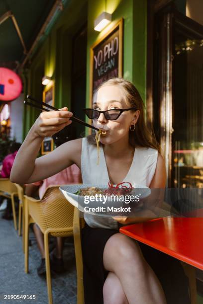 fashionable young woman eating pho while sitting on chair at restaurant - comer fotografías e imágenes de stock