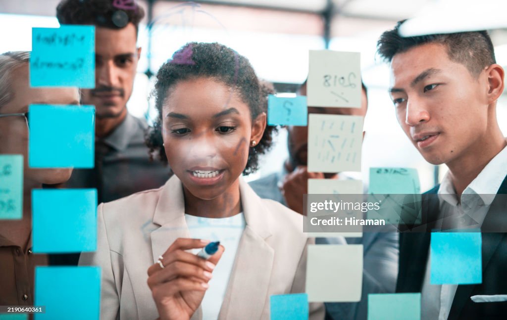 Vielfältiges Team, das an einer transparenten Tafel mit Haftnotizen in einer modernen Büroumgebung zusammenarbeitet
