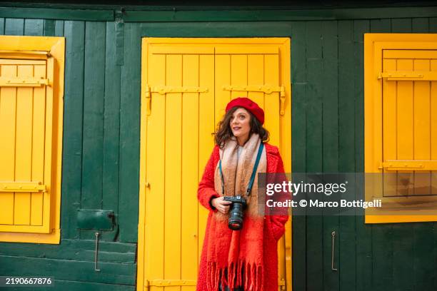 woman in red coat with camera standing by colorful building with yellow shutters, vienna, austria - roter mantel stock-fotos und bilder