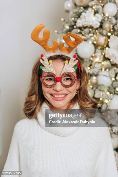 christmas portrait close up woman looking at camera in white sweater wearing christmas glasses and deer antler headband - costume reindeer antlers stock pictures, royalty-free photos & images