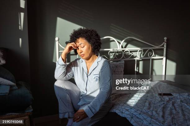 femme en pyjama rayé se sentant stressée alors qu’elle est assise sur le lit dans la lumière du matin - insomnie photos et images de collection