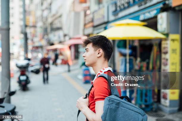young man in a red shirt with a backpack smiles while standing on a bustling city street with shops in the background - korean people stock pictures, royalty-free photos & images