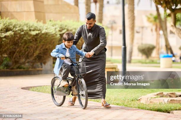 père portant le thawb soutenant son fils faisant du vélo sur le sentier - culture saoudienne photos et images de collection