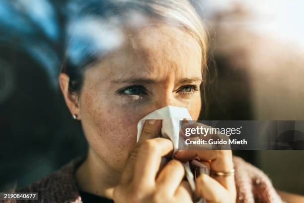 ill woman sneezing into tissue paper while standing behind a window. - allergie photos et images de collection