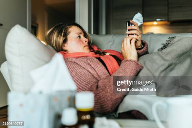 woman lying on a sofa and using her mobile phone writing a sick note while having a flu and feeling sick. - medical condition stock pictures, royalty-free photos & images