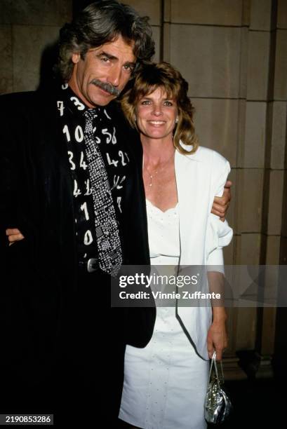 American actor Sam Elliott, wearing a black suit over a black shirt with white number motifs and a black-and-white patterned tie, and his wife,...