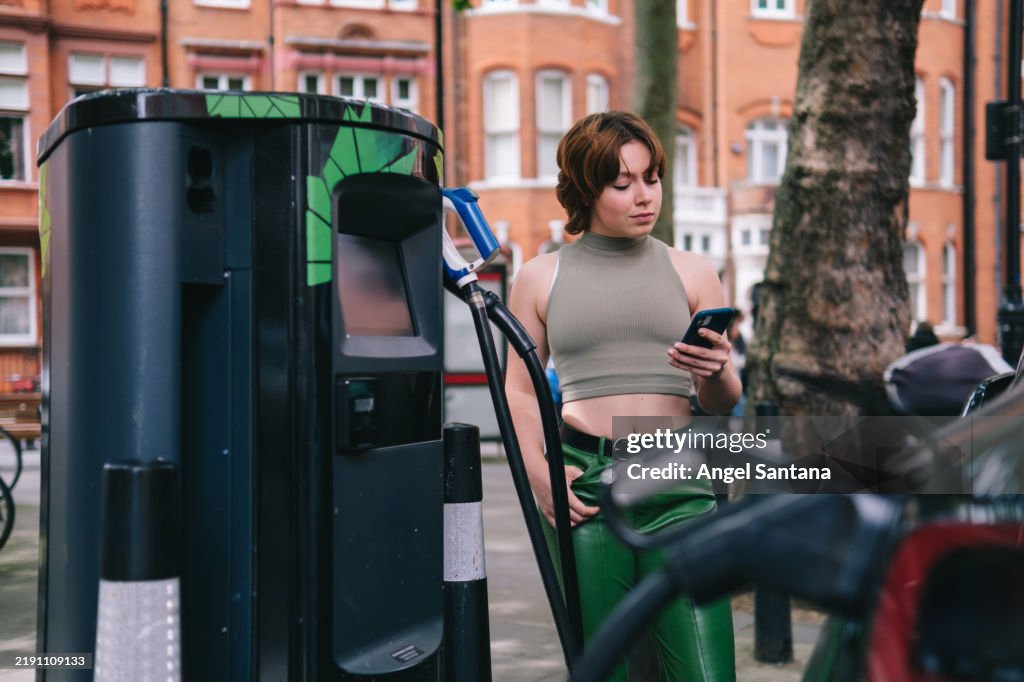 Young woman charging electric car and using her phone