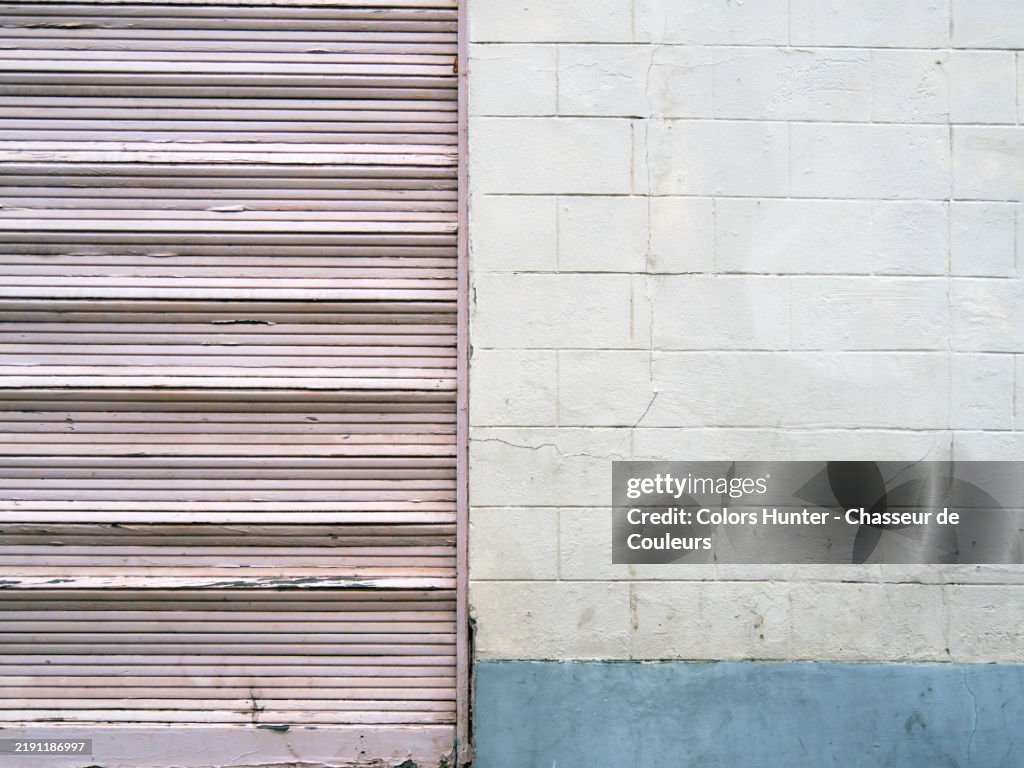 Close-up of a concrete block wall painted white and blue with an old wooden roller shutter painted pink. Natural patina. Brussels, Belgium.