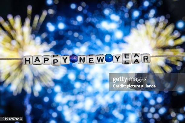 December 2024, North Rhine-Westphalia, Duisburg: ILLUSTRATION - The words "Happy New Year" made of letter cubes hang in front of a photo of fireworks...