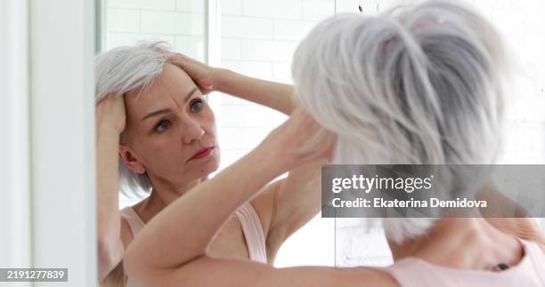 mature woman examining her face and hair in the mirror - grijs haar stockfoto's en -beelden