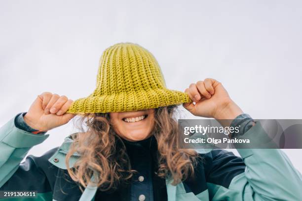 playful woman smiling and covering her face with a woolen hat - yellow green eyes photos et images de collection