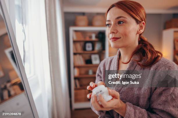 woman wearing bathrobe holding face cream looking in the mirror in the bedroom - vanity mirror stock pictures, royalty-free photos & images