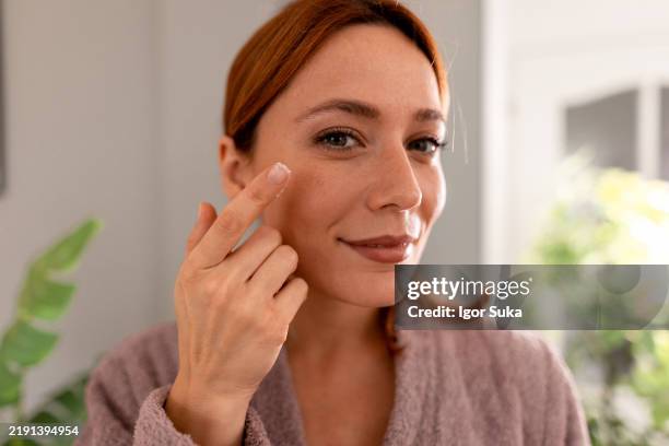 lächelnde frau, die feuchtigkeitscreme auf das gesicht aufträgt und in die kamera schaut - faltenreduktion stock-fotos und bilder