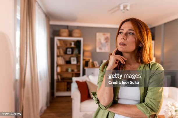 pensive woman thinking with hand on chin in living room - incerteza imagens e fotografias de stock