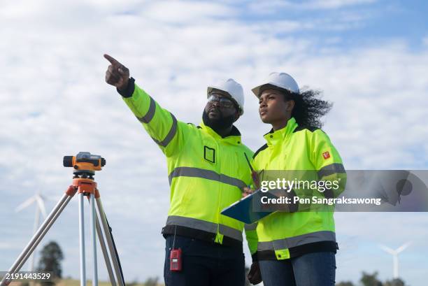 young african american maintenance engineer and woman taking notes and looking through the survey camera to level the area for road construction in a wind power production area. - geodesy stock pictures, royalty-free photos & images