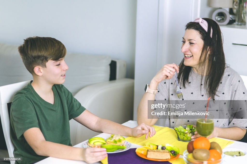 Mother and son enjoying in preparation of a healthy breakfast in the morning at home. Superfoods ingredients for first meal of the day spirulina, smoked tofu, avocado, lettuce, rice cakes, fruits.