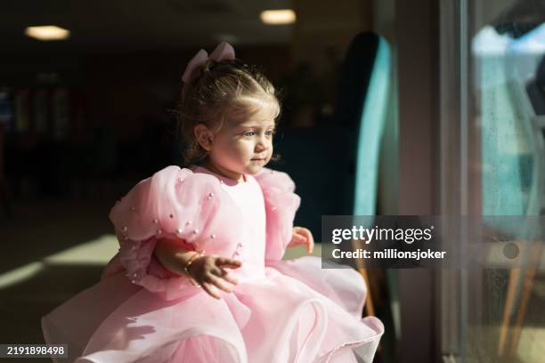 a blonde, blue-eyed little girl looking at her reflection by the window - baby girls stock pictures, royalty-free photos & images